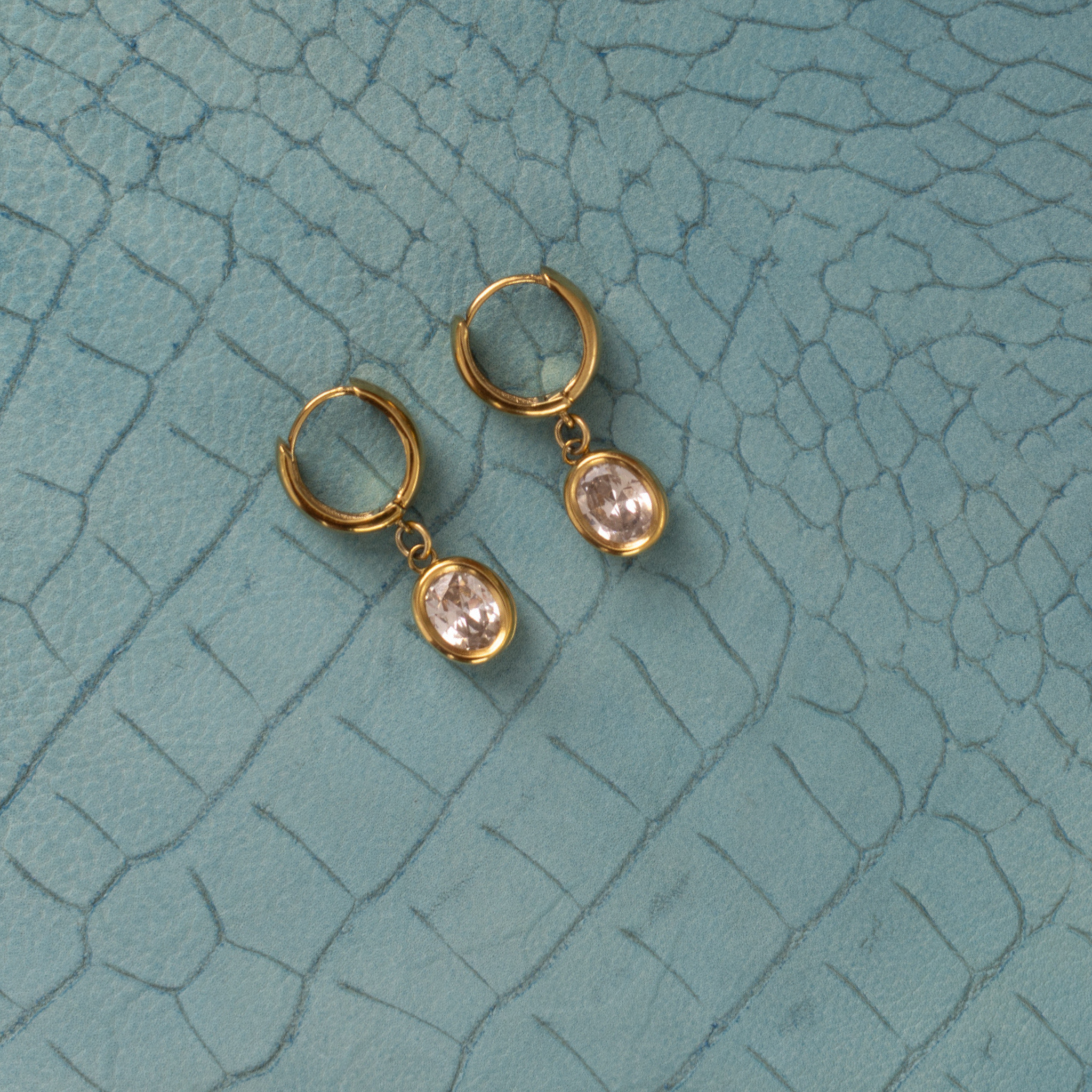 Lanamara Vintage Clarity oval CZ huggie earrings, flat lay on teal