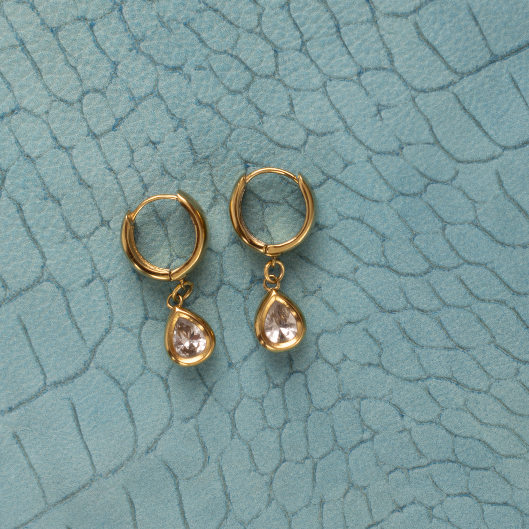 Lanamara Vintage Clarity pear CZ huggie earrings, flat lay on teal